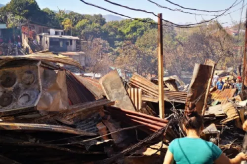 Restos de latas y madera quemadas con una mujer con camisa verde observando la situación